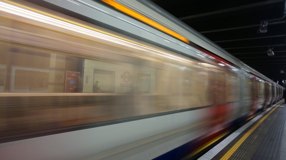 A moving train at a London underground station, captured in motion blur. The train's exterior has a metallic surface with horizontal panels and a streak of yellow along the top, indicating the tube's 
